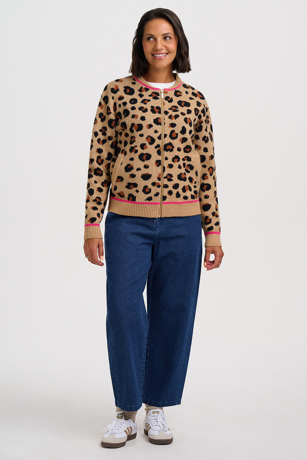 A woman modeling a beige leopard-print zip-up cardigan with pink and beige ribbed trim, paired with blue jeans and white sneakers, against a light gray studio background.