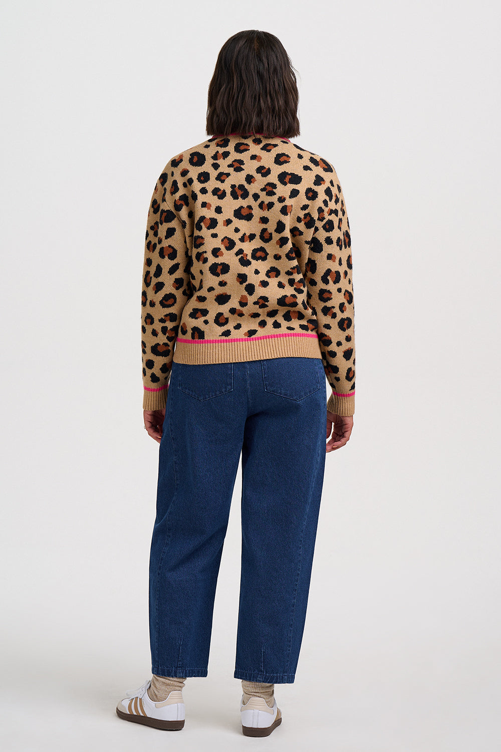 A model shown from the back wearing a beige leopard-print knit sweater with a pink ribbed hem and cuffs, paired with dark blue jeans and white sneakers, against a plain light gray studio background.