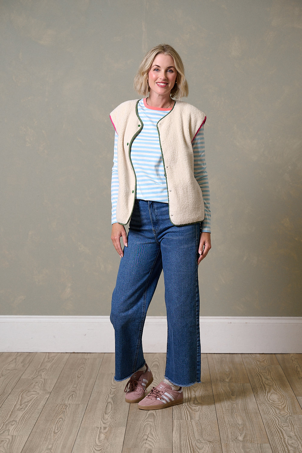 Woman wearing a cream vest, striped shirt, and blue jeans against a plain wall.