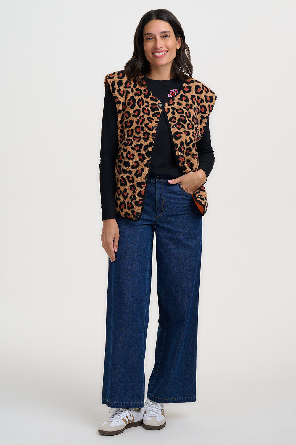 Woman wearing a leopard print vest, black top, blue jeans, and white sneakers on a white background