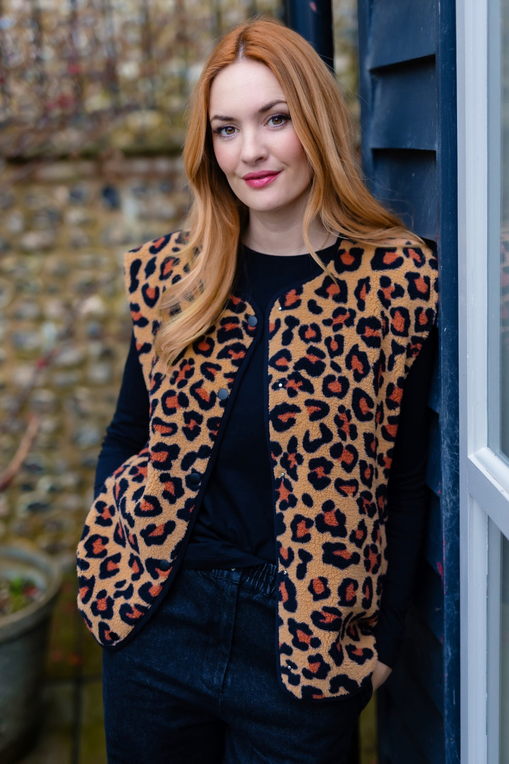 A woman with long red hair wearing a fuzzy leopard-print vest over a black top and dark jeans, standing outdoors next to a dark blue door.
