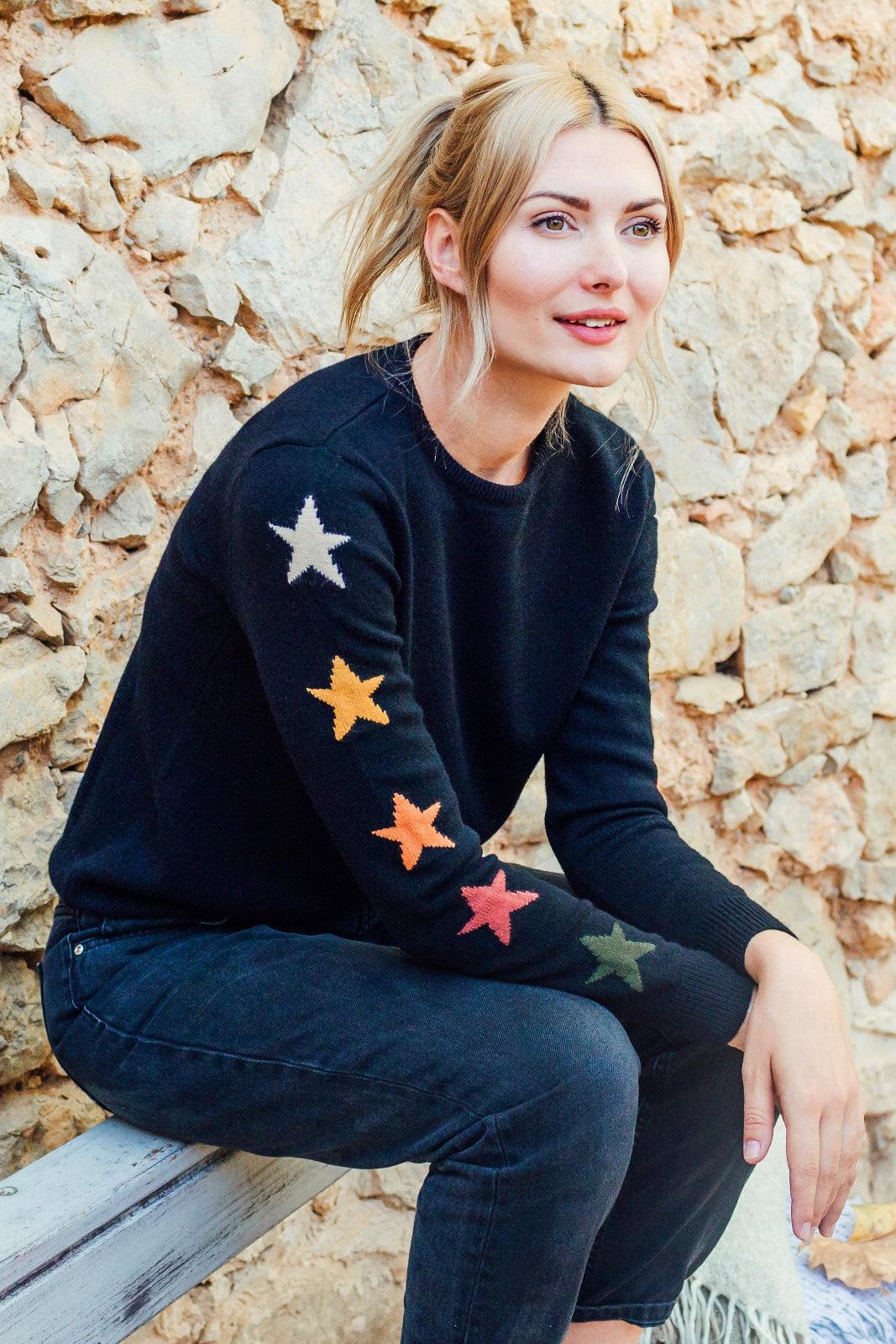 A woman sitting against a stone wall, wearing a black crew neck jumper with multicolored star patterns on the sleeves.