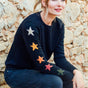 A woman sitting against a stone wall, wearing a black crew neck jumper with multicolored star patterns on the sleeves.