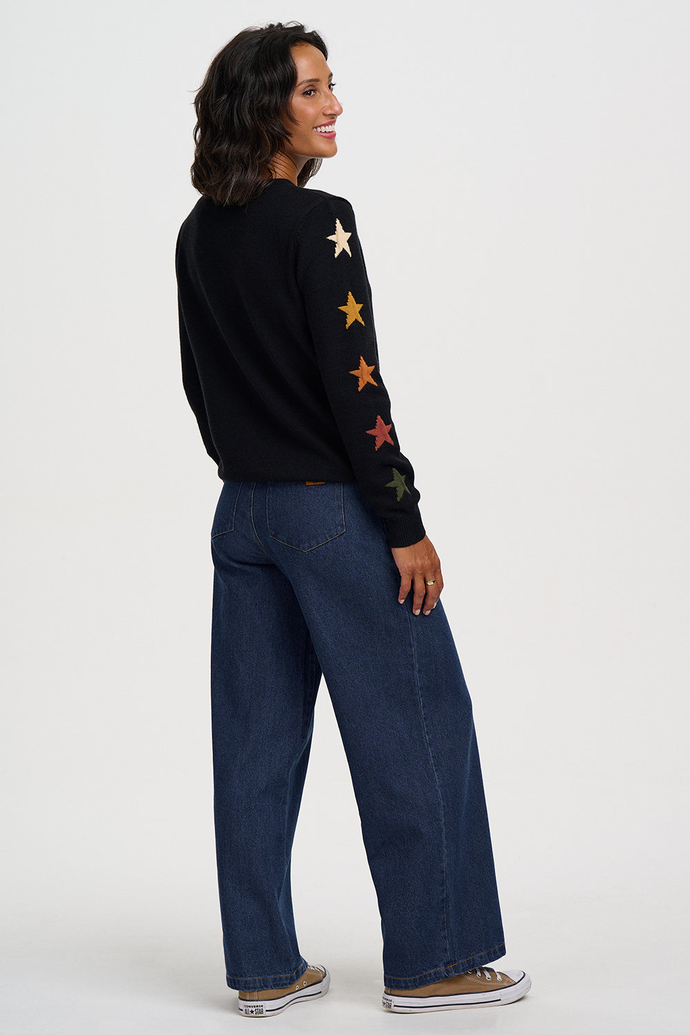 A woman shown from the back wearing a black knit sweater with colorful embroidered stars down one sleeve, paired with wide-leg dark blue jeans and brown sneakers, against a plain light gray studio background.