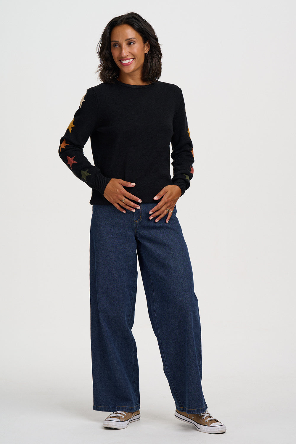 A woman modeling a black long-sleeve knit top with colorful embroidered star patterns on the sleeves, paired with dark blue wide-leg jeans and brown sneakers, against a light gray studio background.