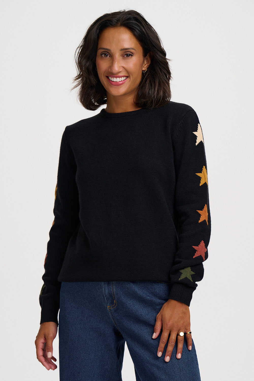 A woman wearing a black crewneck sweater with colorful embroidered stars on the sleeves, paired with blue jeans, against a plain light gray background.