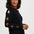 A woman wearing a black crewneck sweater with colorful embroidered stars on the sleeves, paired with blue jeans, photographed against a light gray studio background.