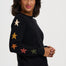 A woman wearing a black crewneck sweater with colorful embroidered stars on the sleeves, paired with blue jeans, photographed against a light gray studio background.