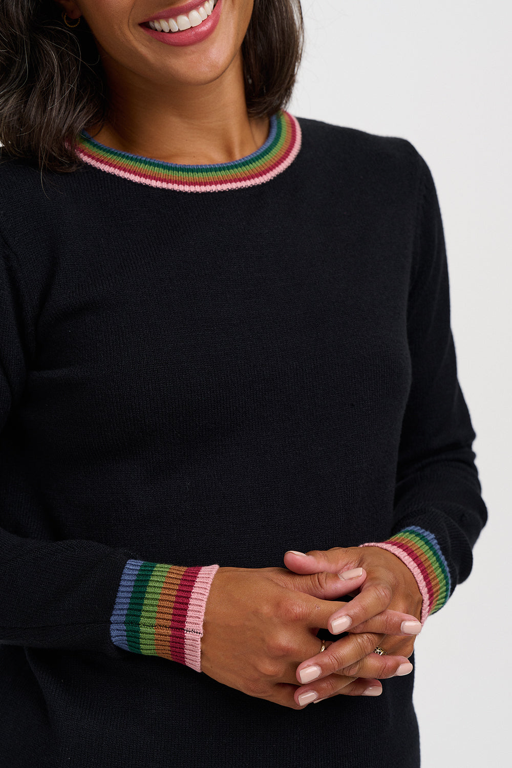 A close-up of a woman wearing a black crewneck sweater with colorful striped ribbed trim at the collar and cuffs. The sweater is shown against a plain light gray background.