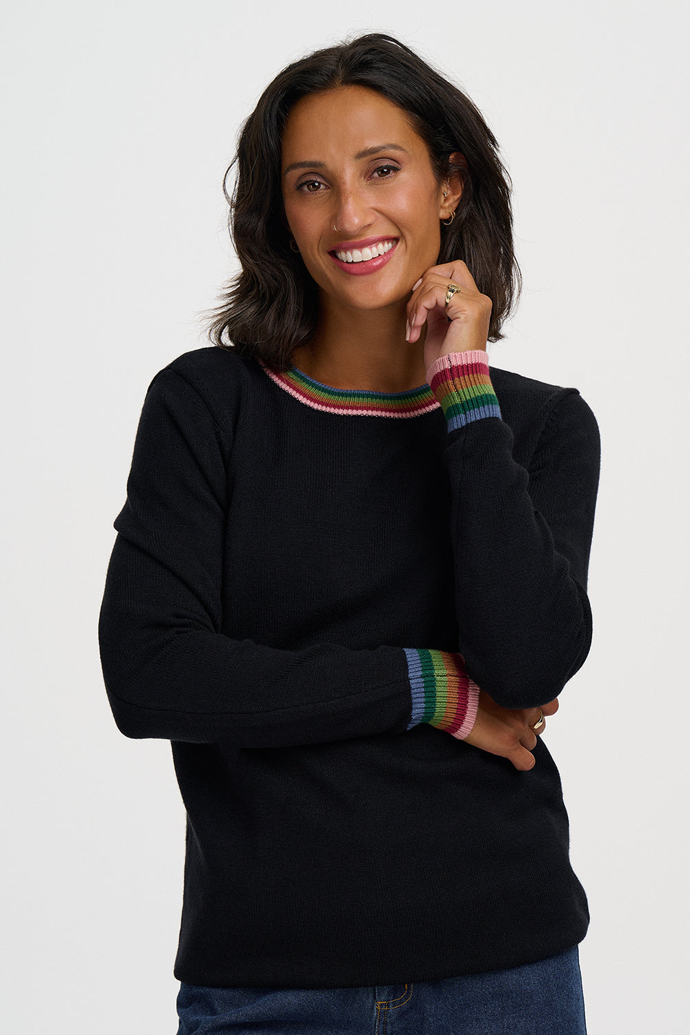 A woman wearing a black crewneck sweater with rainbow-striped trim at the collar and cuffs, paired with blue jeans, photographed against a light gray studio background.