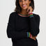 A woman wearing a black crewneck sweater with rainbow-striped trim at the collar and cuffs, paired with blue jeans, photographed against a light gray studio background.