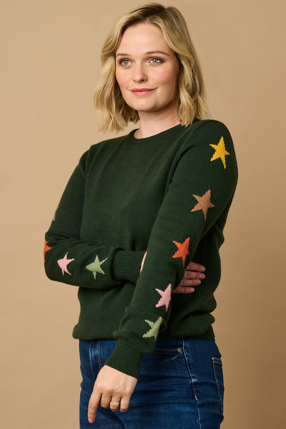 A woman wearing a green crew neck jumper with multicolored star intarsia sleeves, paired with blue jeans.
