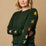 A woman wearing a green crew neck jumper with multicolored star intarsia sleeves, paired with blue jeans.