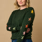 A woman wearing a green crew neck jumper with multicolored star intarsia sleeves, paired with blue jeans.