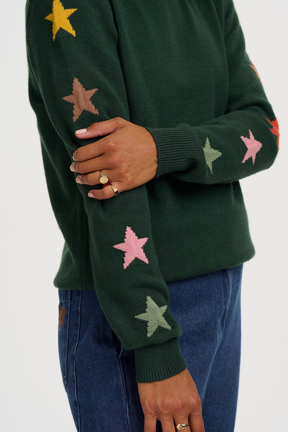 A close-up of a person wearing a dark green knit sweater with colorful embroidered star patches in pink, yellow, and light green, paired with blue jeans.