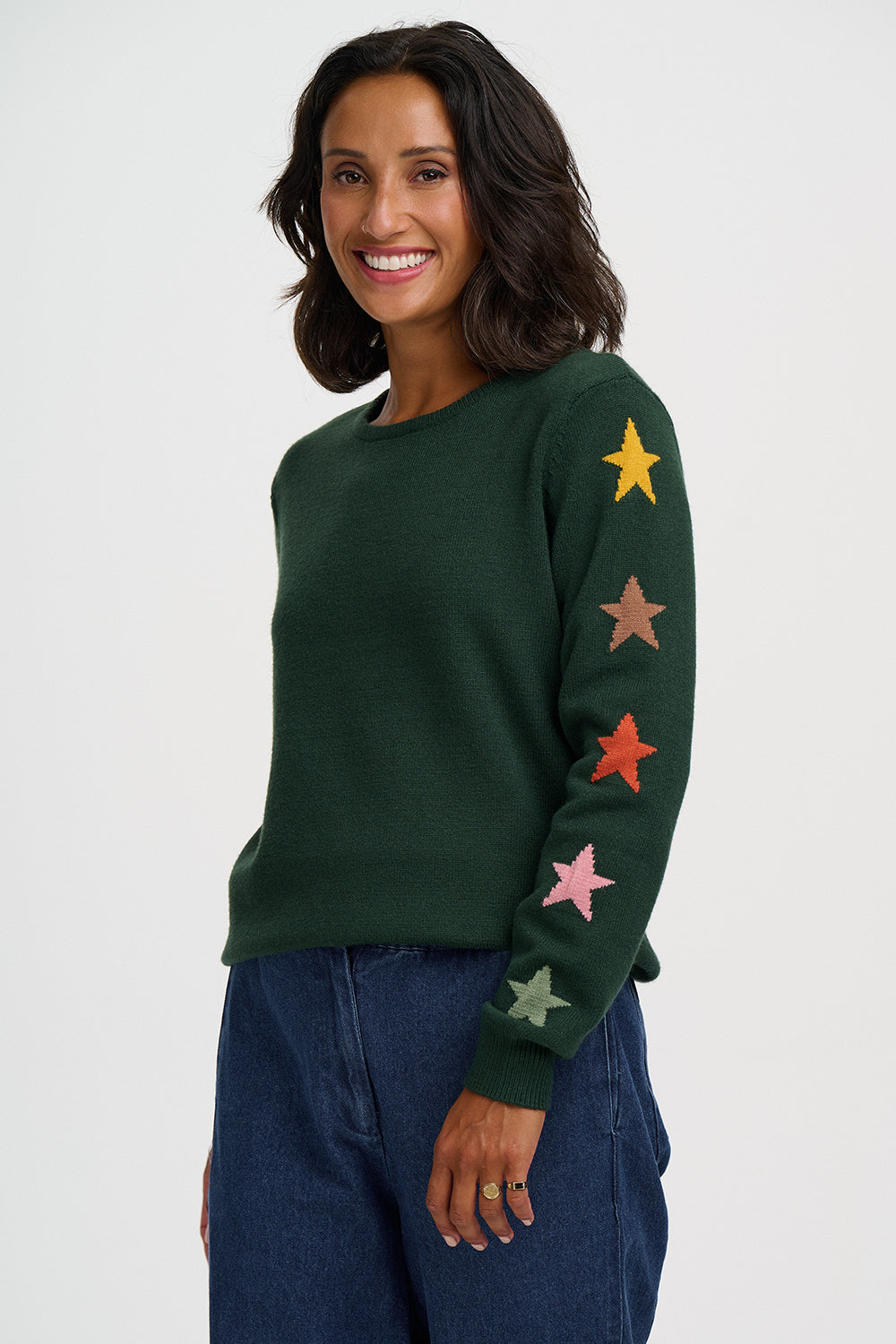 A woman wearing a dark green crewneck sweater with colorful embroidered stars down one sleeve, paired with dark blue jeans, against a plain light gray background.