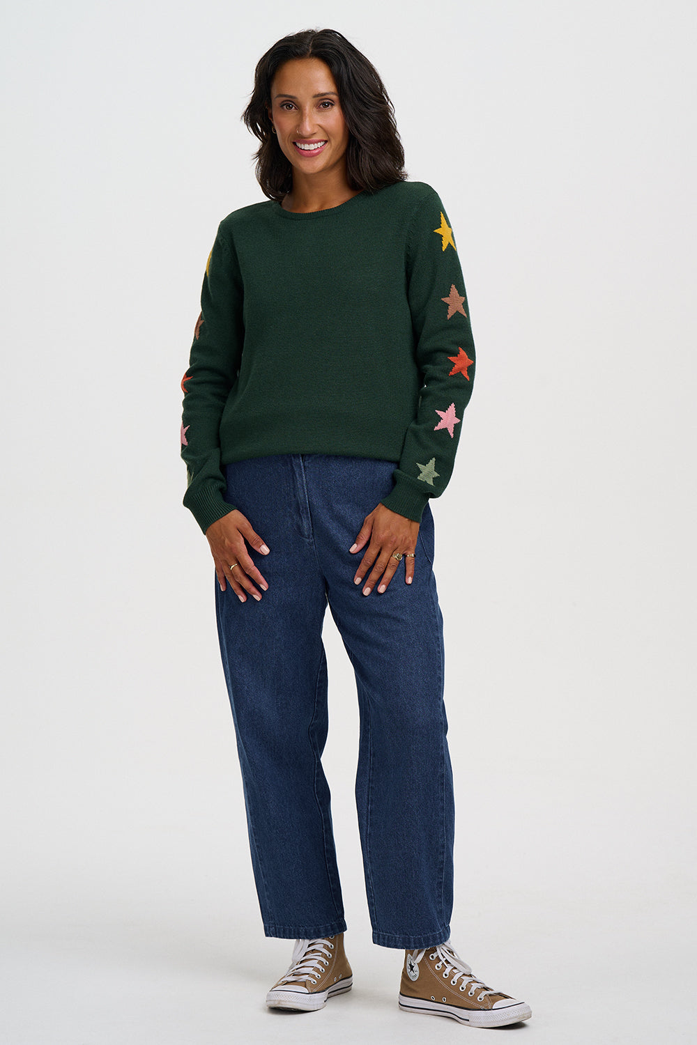 A woman modeling a dark green crewneck sweater with colorful embroidered stars on the sleeves, paired with dark blue jeans and brown sneakers, against a light gray studio background.