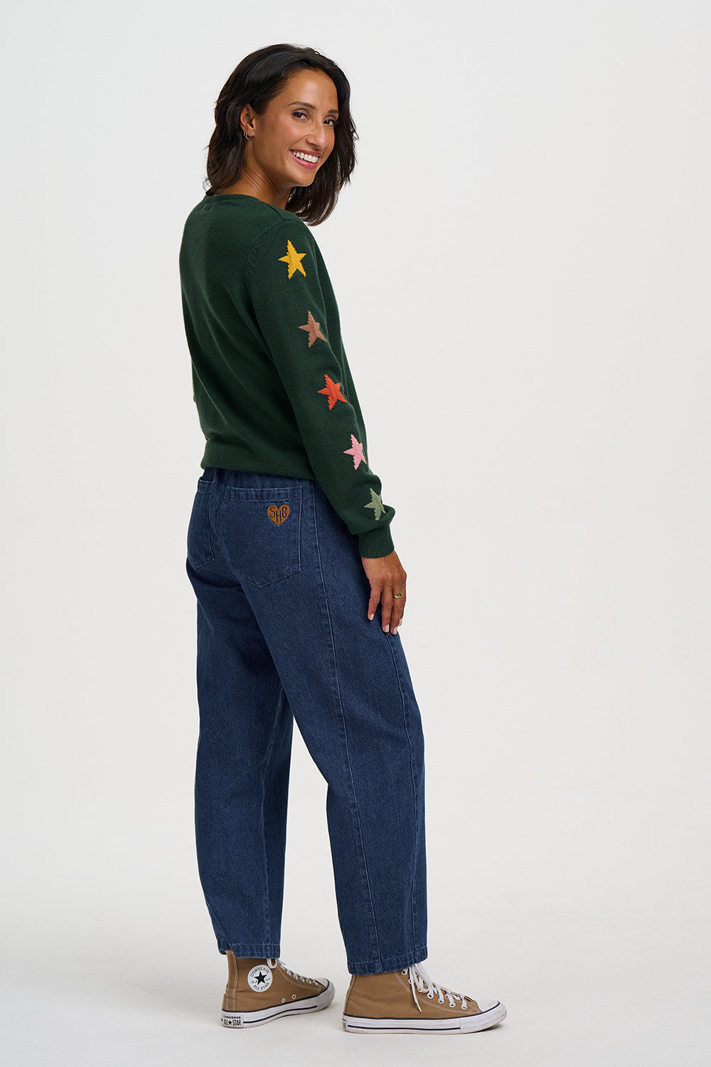 A woman modeling a dark green crewneck sweater with colorful embroidered stars down the sleeves, paired with dark blue jeans and brown Converse sneakers, shown from a side-back angle against a light gray studio background.