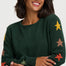 A woman wearing a dark green crewneck sweater with colorful star patterns in yellow, orange, and green, posed against a light gray background.