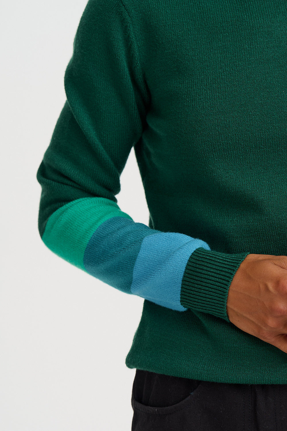 Close-up of a person wearing a green knit sweater with color-blocked elbow patches in teal and light blue, plus a dark ribbed cuff, against a light gray background.
