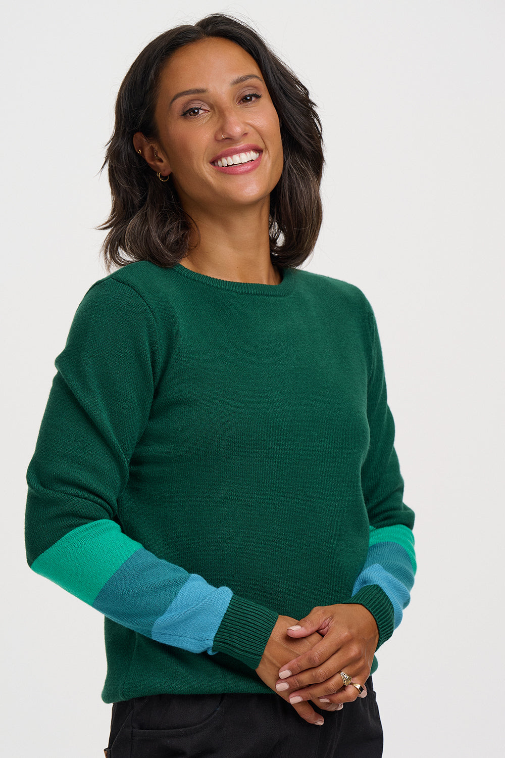 A smiling woman wearing a dark green crewneck sweater with contrasting teal and light blue striped sleeves, posed against a solid light gray background.