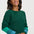 A smiling woman wearing a dark green crewneck sweater with contrasting teal and light blue striped sleeves, posed against a solid light gray background.