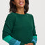 A smiling woman wearing a dark green crewneck sweater with contrasting teal and light blue striped sleeves, posed against a solid light gray background.