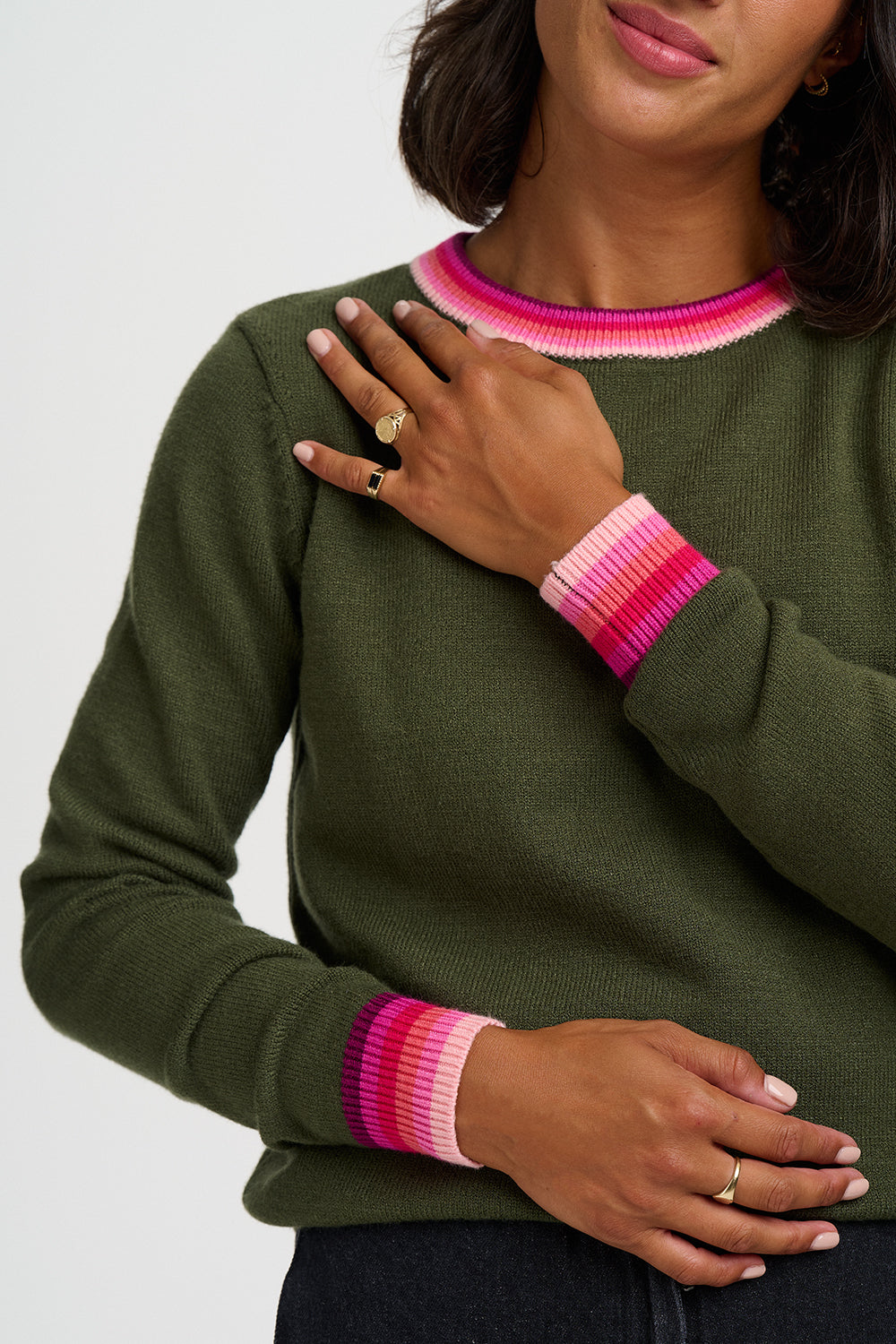 An olive green crewneck sweater with vibrant pink and magenta striped ribbed trim at the collar and cuffs, shown on a model against a light gray background.