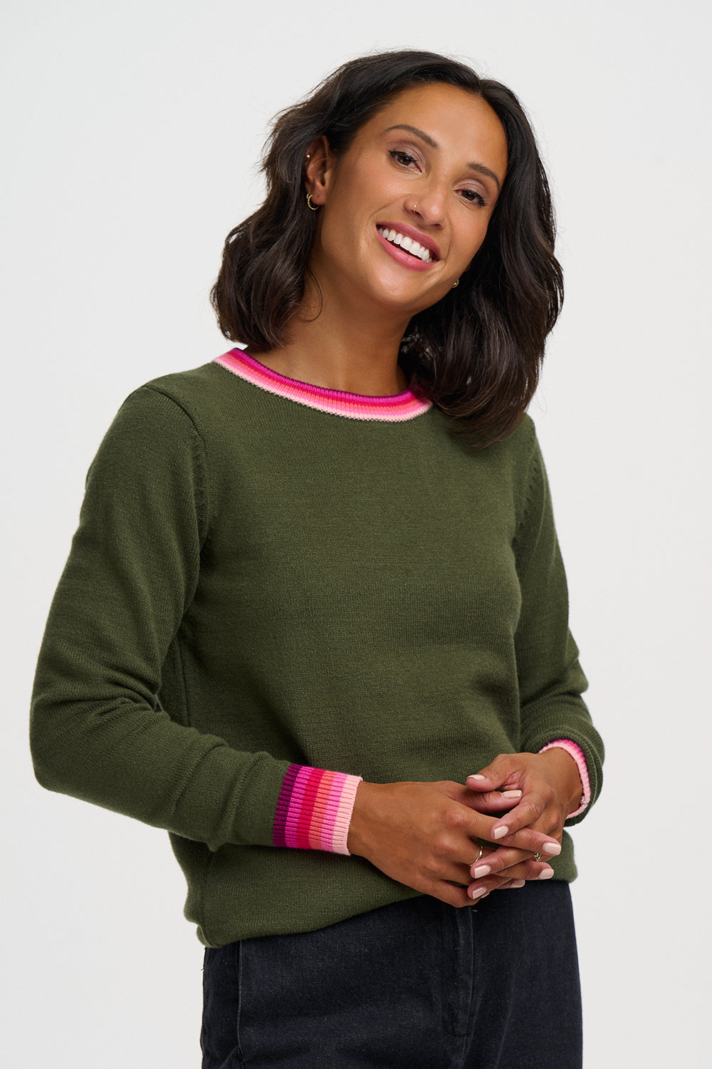 A woman wearing an olive green crewneck sweater with pink and white striped ribbed trim at the collar and cuffs, paired with dark jeans against a light gray studio background.