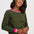 A woman wearing an olive green crewneck sweater with pink and white striped ribbed trim at the collar and cuffs, paired with dark jeans against a light gray studio background.
