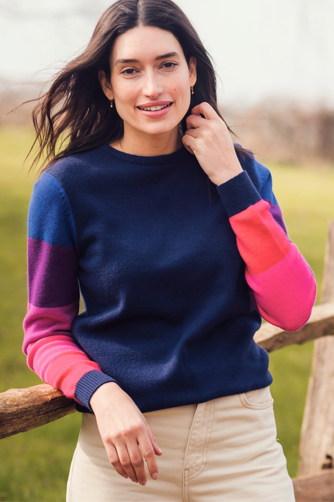 A woman wearing a navy crew neck jumper with color block sleeves in pink and purple, paired with beige pants, standing outdoors.