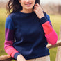 A woman wearing a navy crew neck jumper with color block sleeves in pink and purple, paired with beige pants, standing outdoors.