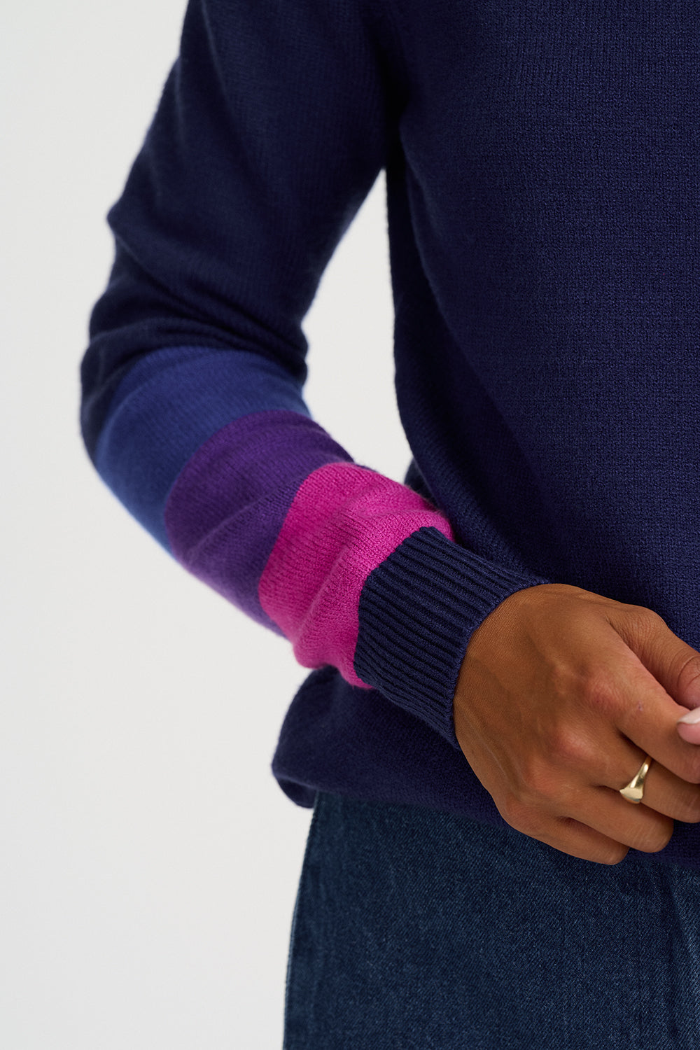Close-up of a person wearing a navy knit sweater with color-blocked cuffs in purple and bright pink, paired with a dark blue skirt or trousers against a light gray studio background.