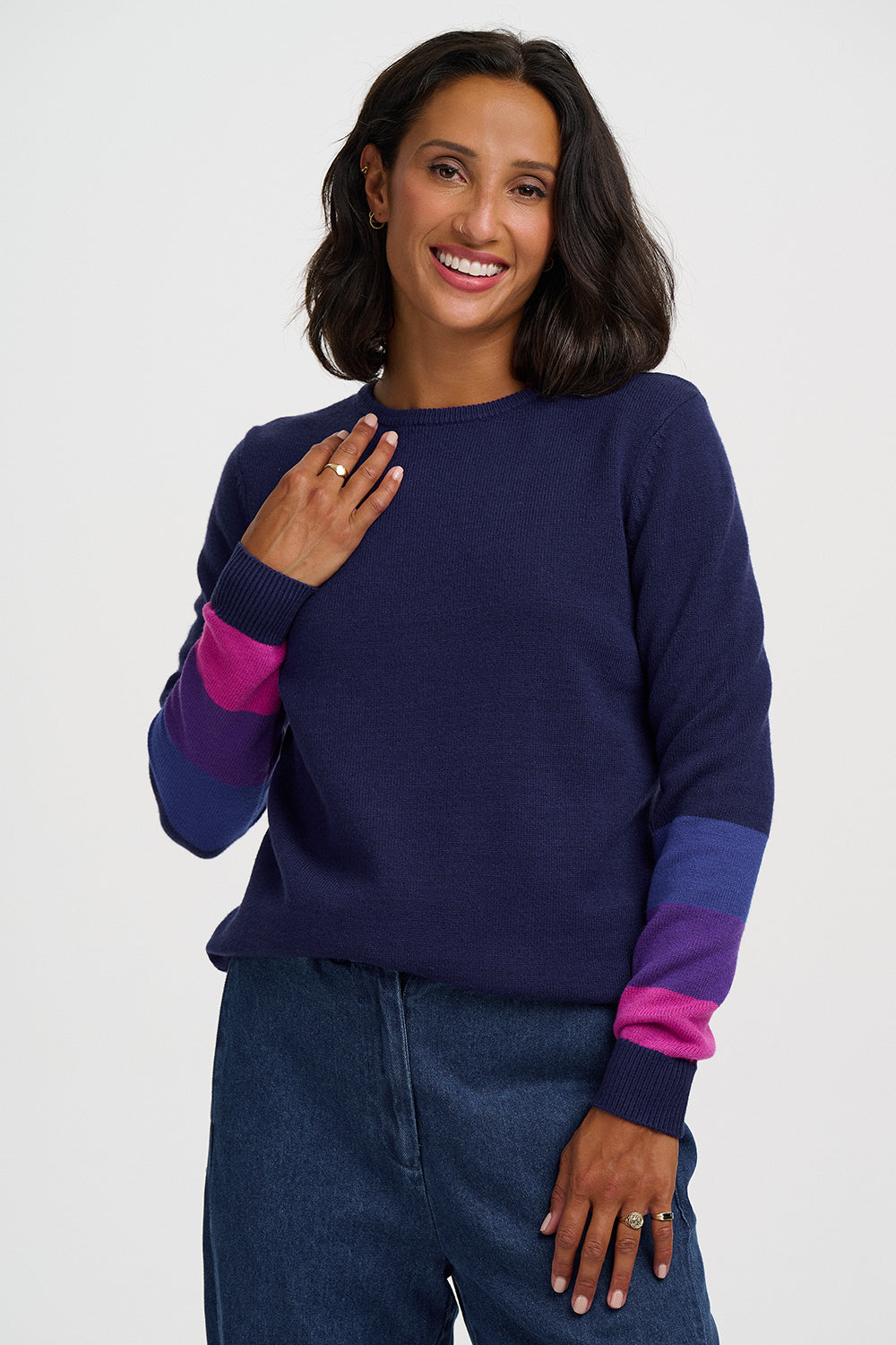 A woman wearing a navy blue knit sweater with color-blocked purple and pink cuffs, paired with dark blue jeans, against a light gray studio background.