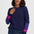 A woman wearing a navy blue knit sweater with color-blocked purple and pink cuffs, paired with dark blue jeans, against a light gray studio background.