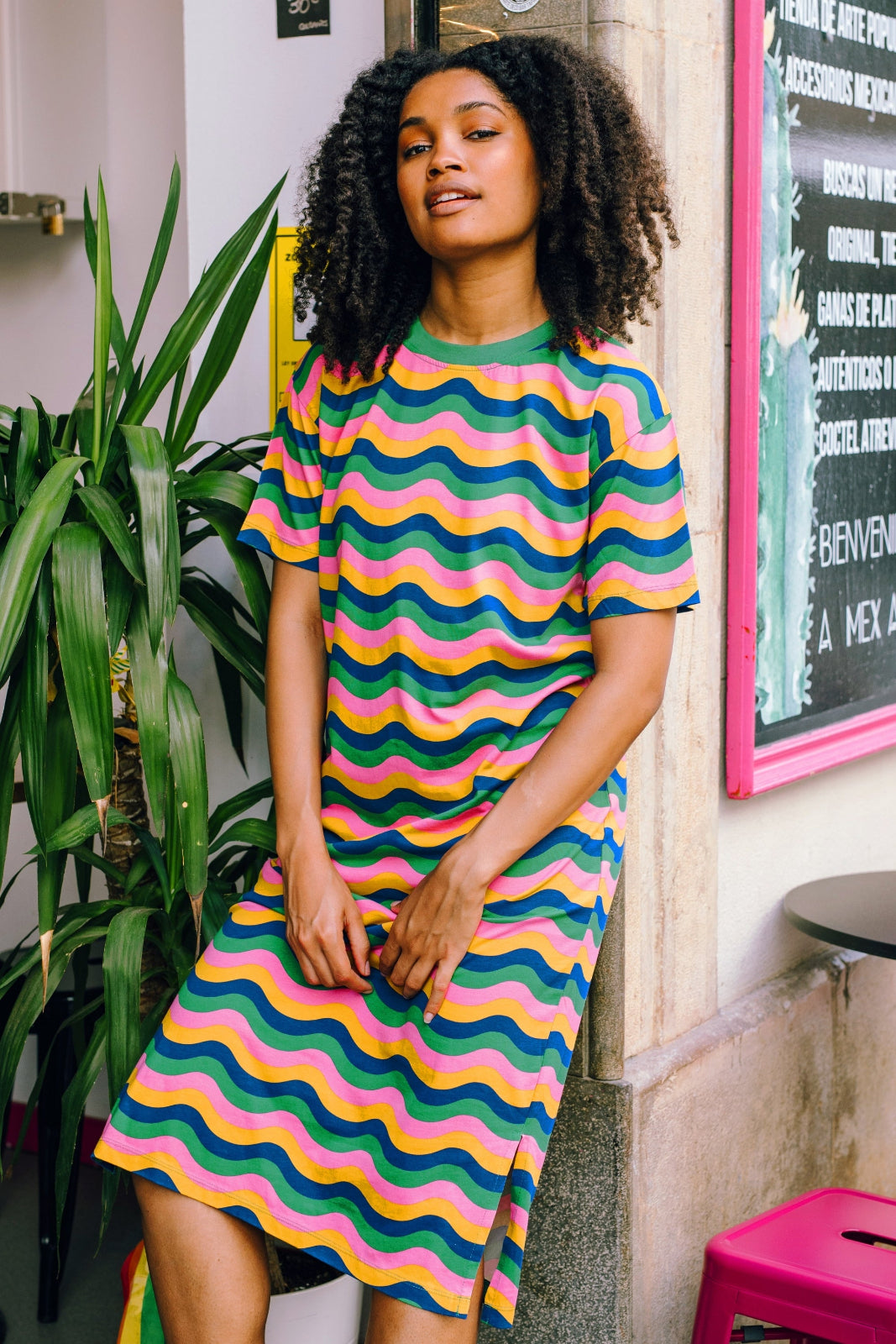 Woman wearing a colorful wavy pattern dress standing indoors with plants and a blackboard in the background.