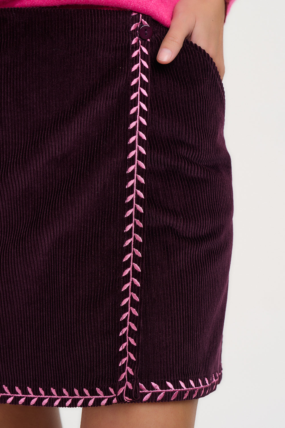 Close-up of a dark purple corduroy skirt with pink contrast stitching, worn by a model whose hand is in a pocket.