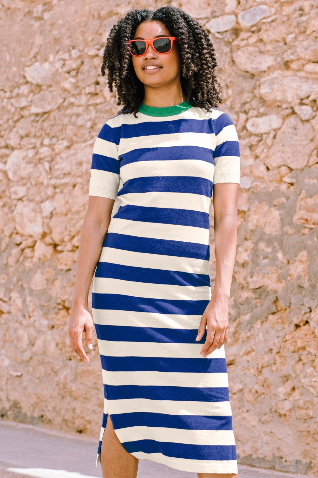 A model wearing a short-sleeved blue and white striped midi knit dress with a contrasting green neckline, standing against a stone wall background.
