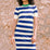 A model wearing a short-sleeved blue and white striped midi knit dress with a contrasting green neckline, standing against a stone wall background.