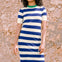 A model wearing a short-sleeved blue and white striped midi knit dress with a contrasting green neckline, standing against a stone wall background.