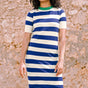 A model wearing a short-sleeved blue and white striped midi knit dress with a contrasting green neckline, standing against a stone wall background.