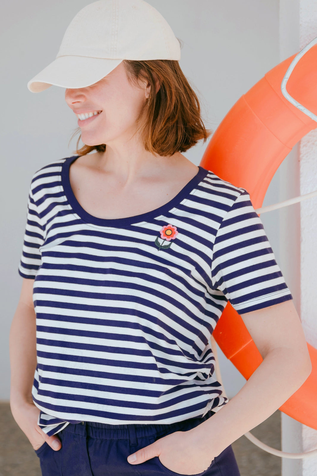 A woman wearing a striped scoop neck t-shirt in white and navy colors with a small floral design on the chest, paired with a white cap and dark shorts.
