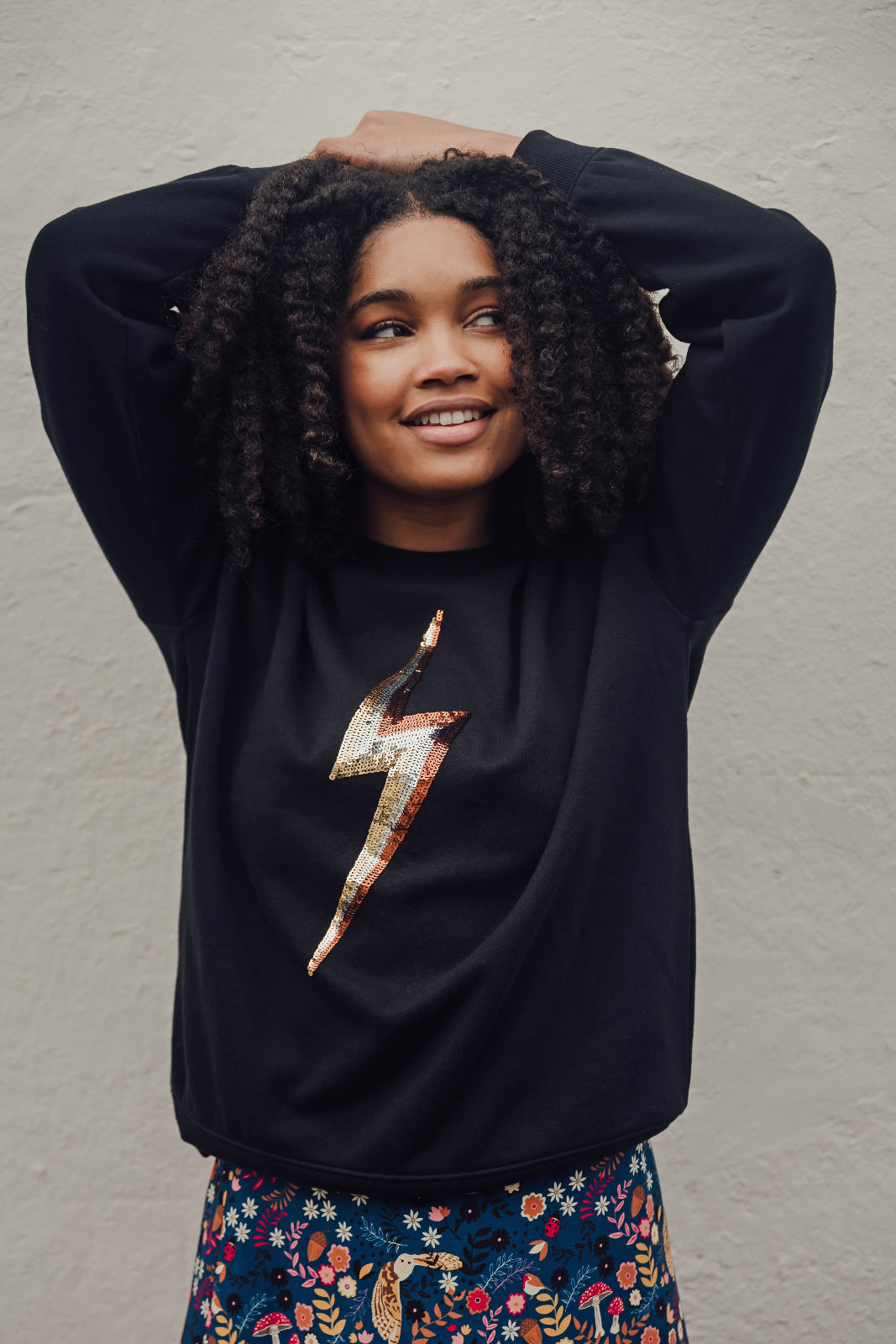 A person wearing a black sweatshirt with a sequin lightning bolt design on the front, posing with one hand raised to their head.