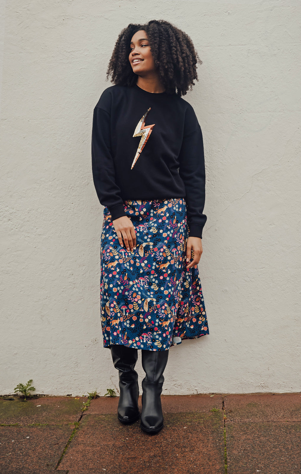 Woman wearing a black sweatshirt with a lightning bolt design, floral skirt, and black boots against a white wall.