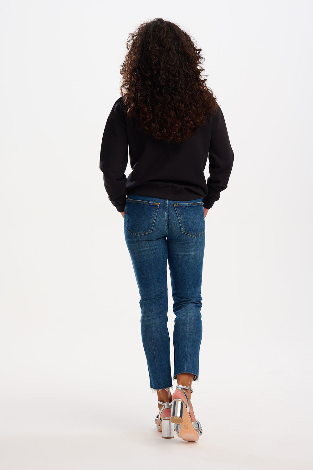 A woman standing, wearing a black sweatshirt with sequin waves in peach, emerald, and blue, and blue jeans.
