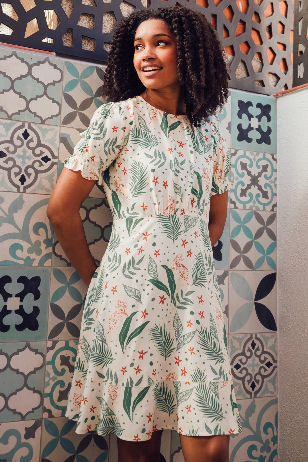 A model wearing a short-sleeved mini dress with a fitted waist and a frill hem skirt, featuring a jungle leaves print on a cream base.