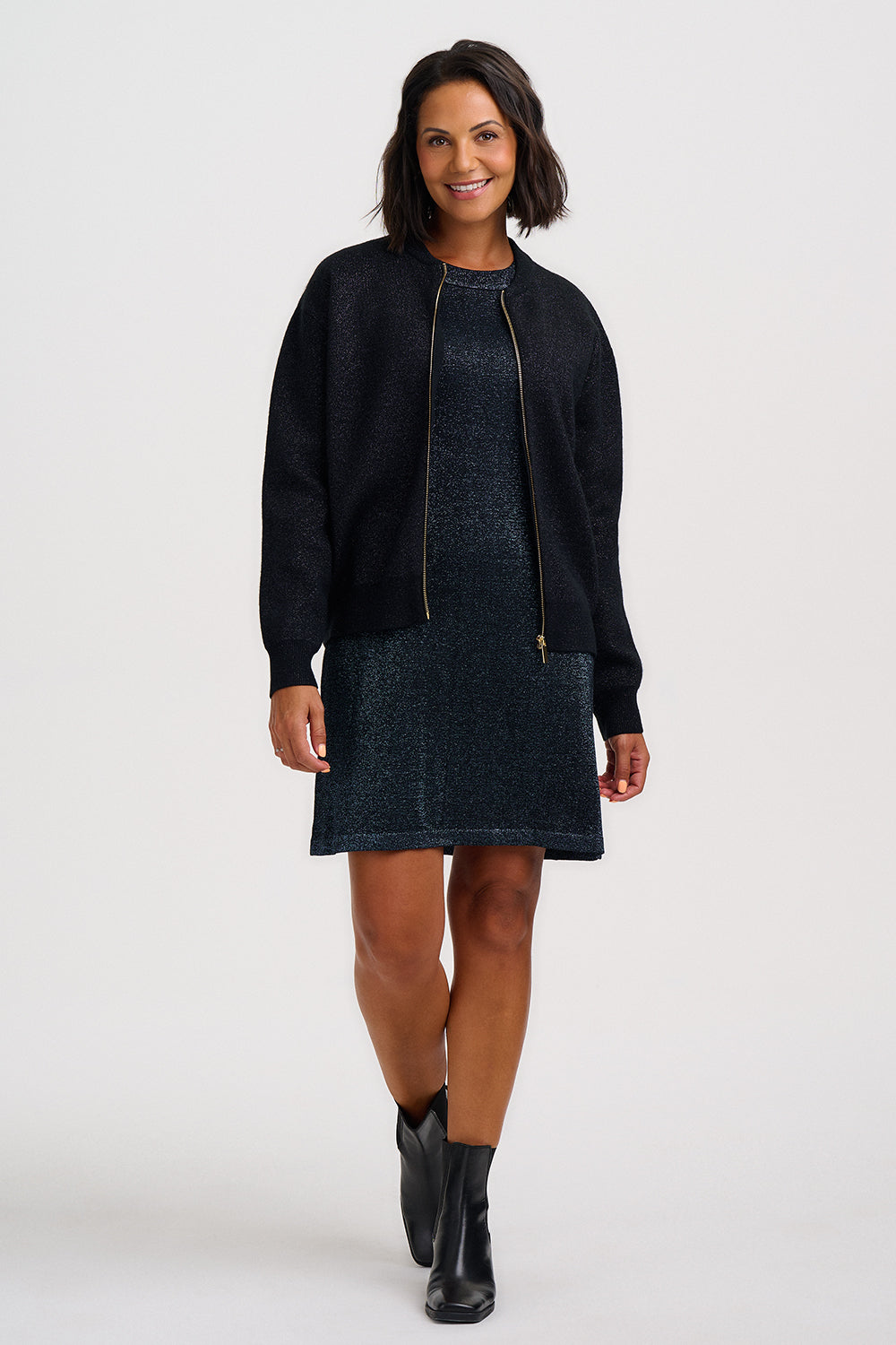 A woman modeling a dark navy textured knit cardigan with gold-tone zippers over a matching dark navy knit dress, paired with black ankle boots, against a plain light gray studio background.