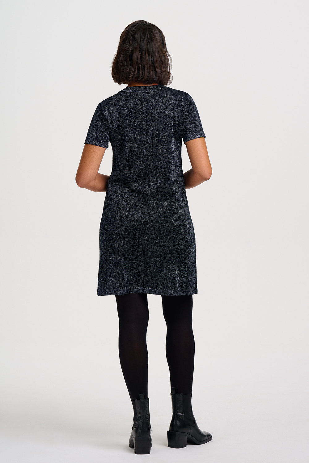 A woman viewed from the back wearing a short-sleeve textured dark gray tunic dress, black tights, and black ankle boots against a plain light gray studio background.