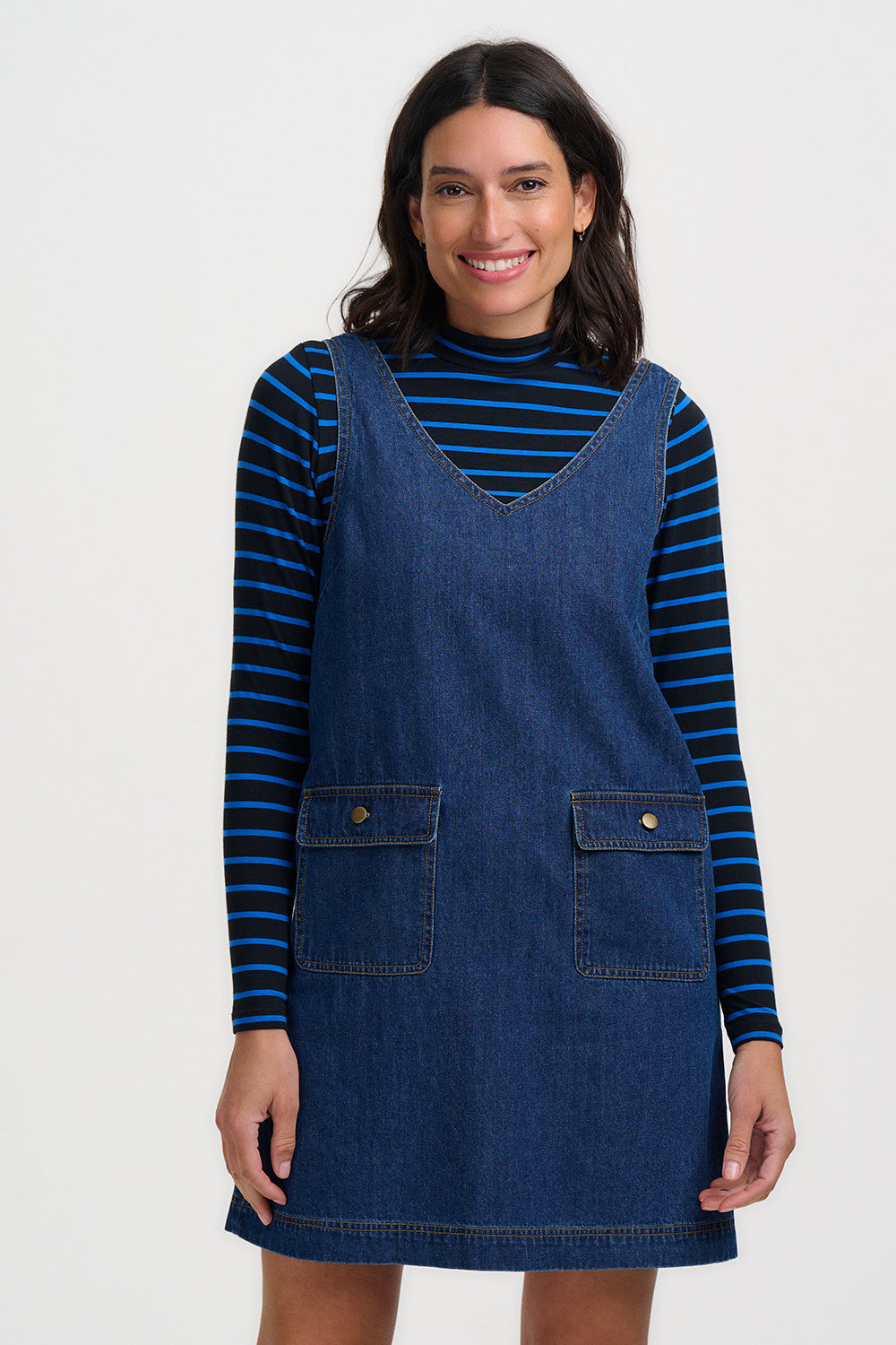 Woman wearing a blue denim pinafore dress over a striped shirt on a white background
