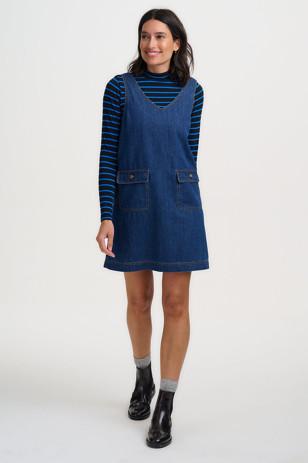 Woman wearing a blue denim pinafore dress over a striped shirt on a white background